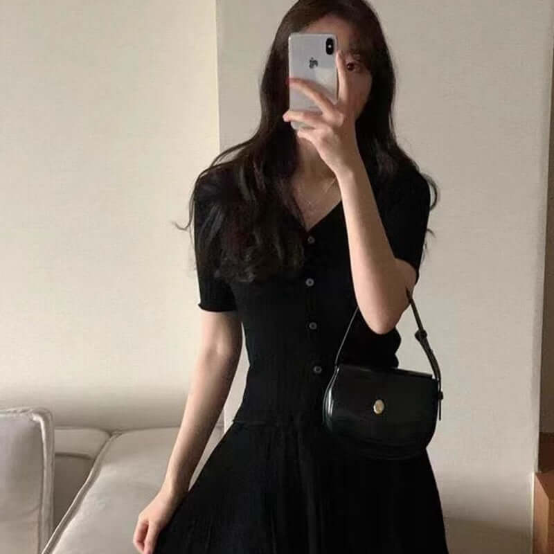 Stylish young woman in a black button-up dress, holding a small black handbag, posing in a modern indoor setting.