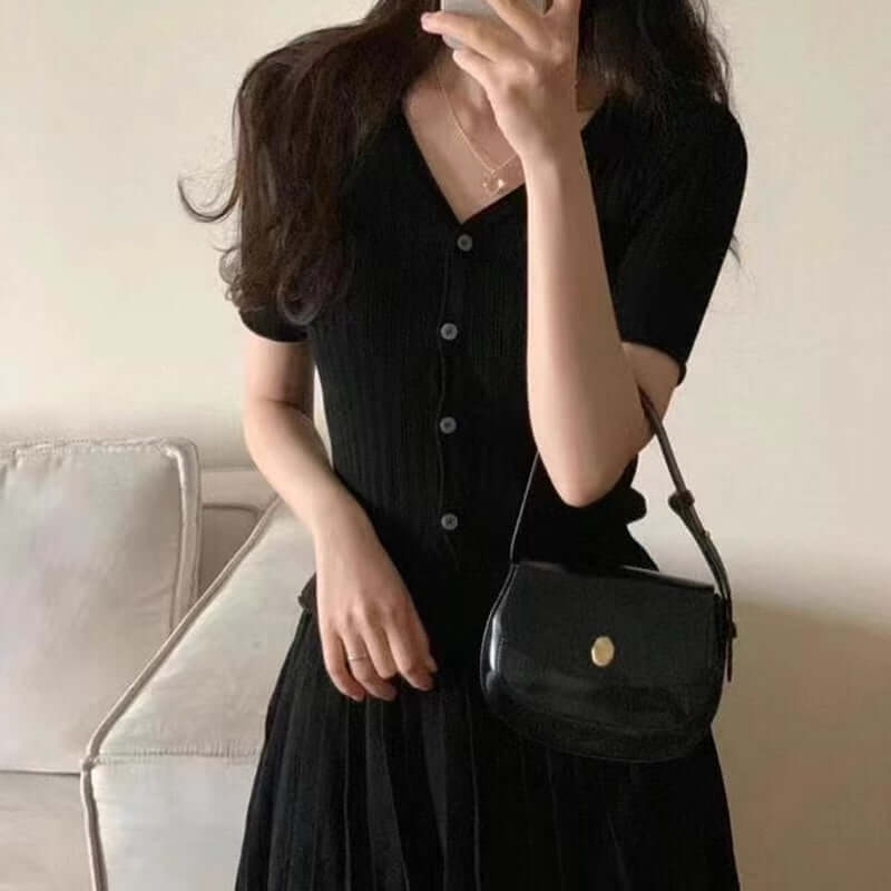 Model wearing a black pleated dress with a button front and a small black handbag in a cozy room.