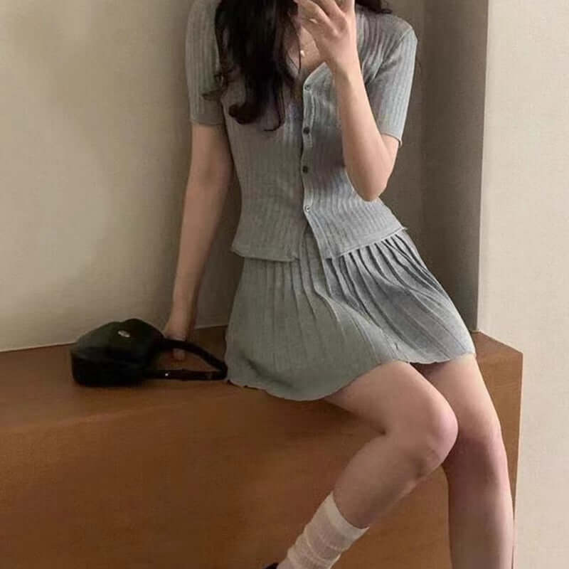 A young woman wearing a chic, gray pleated dress with a fitted top, seated on a wooden surface.